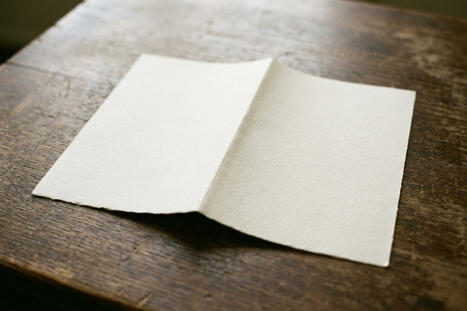 A single folded sheet of pale blank paper lying open on a bare wooden table