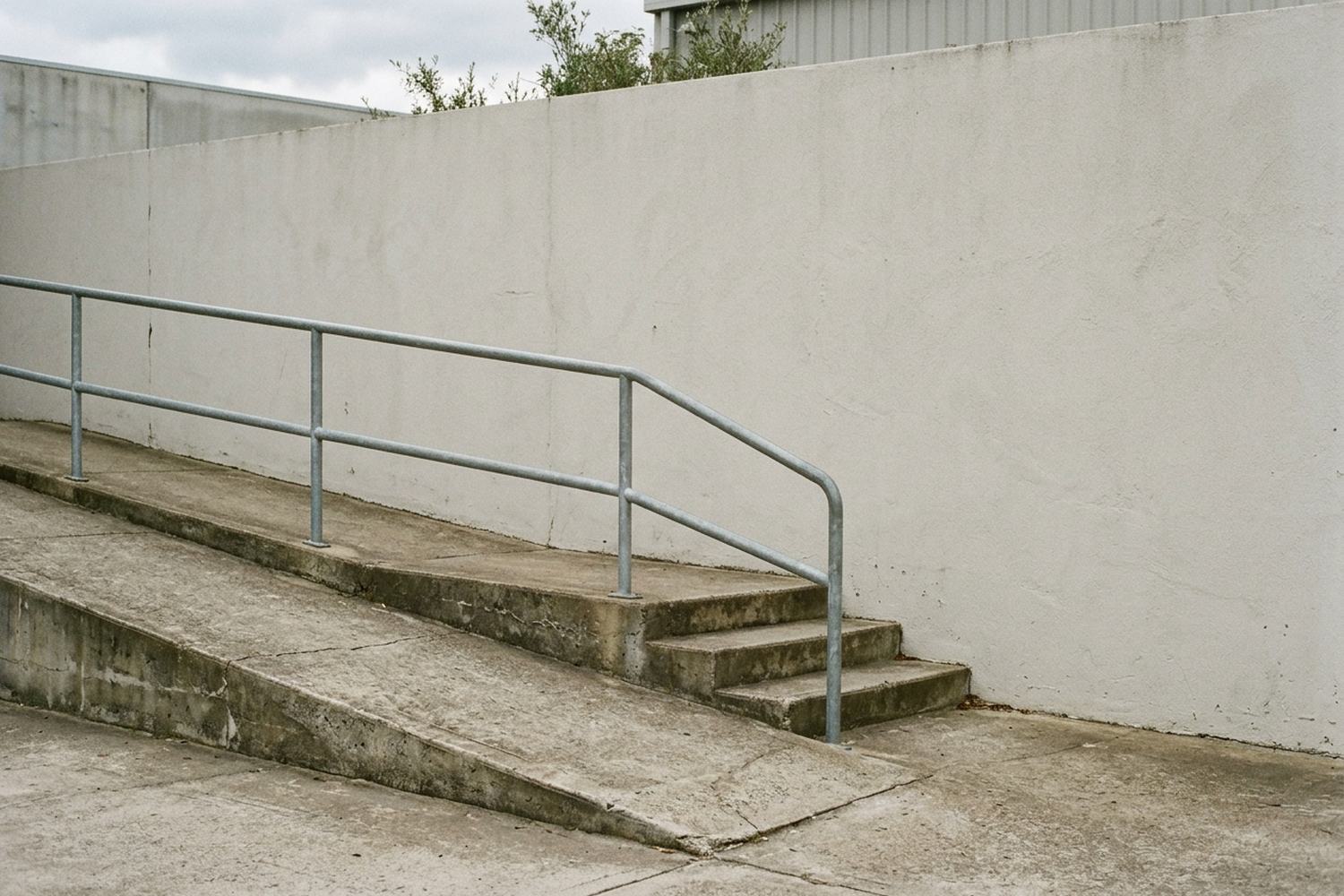 A concrete ramp climbing beside three shallow steps with a plain metal handrail along a rendered wall