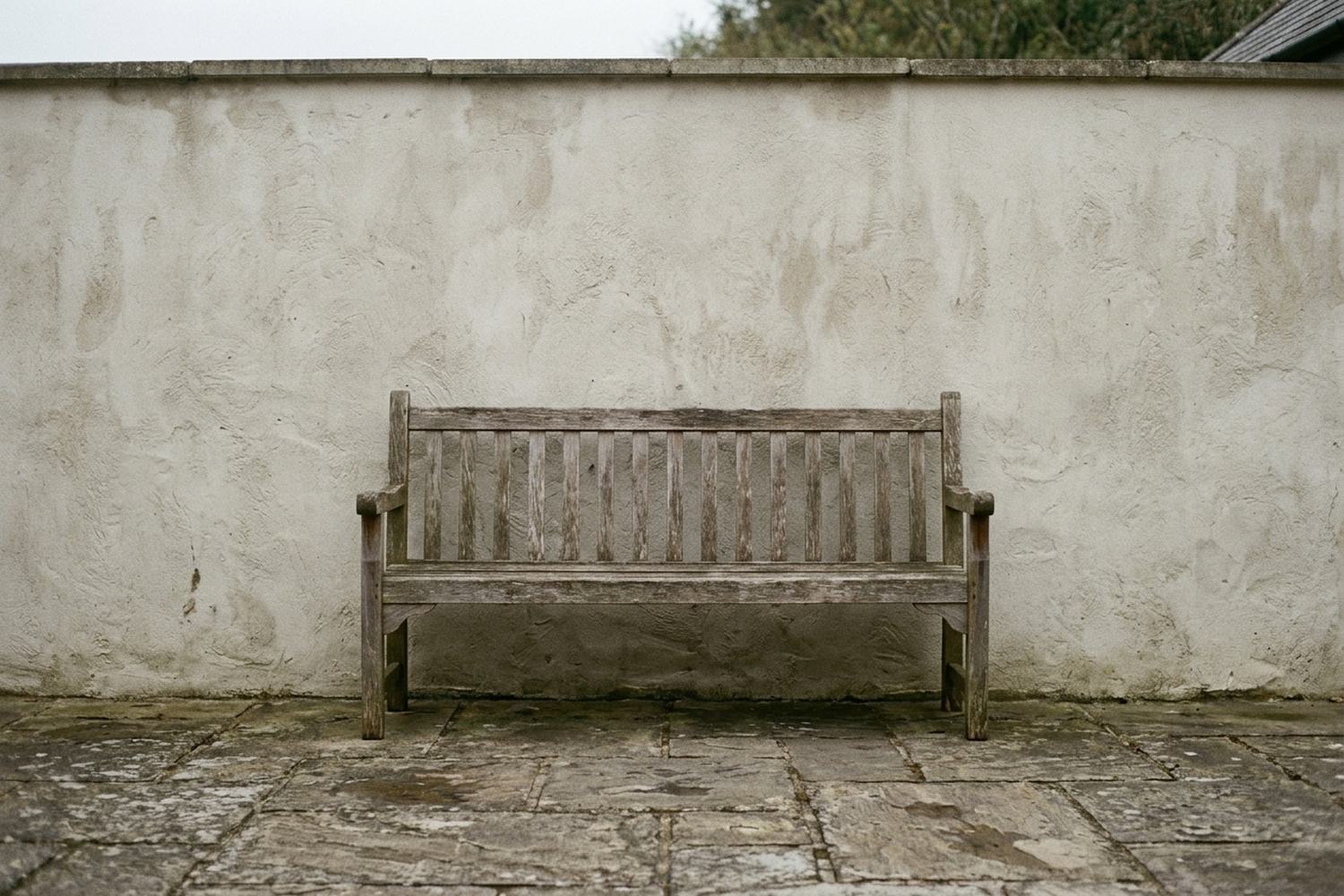 A weathered wooden bench standing empty against a bare rendered wall on worn paving