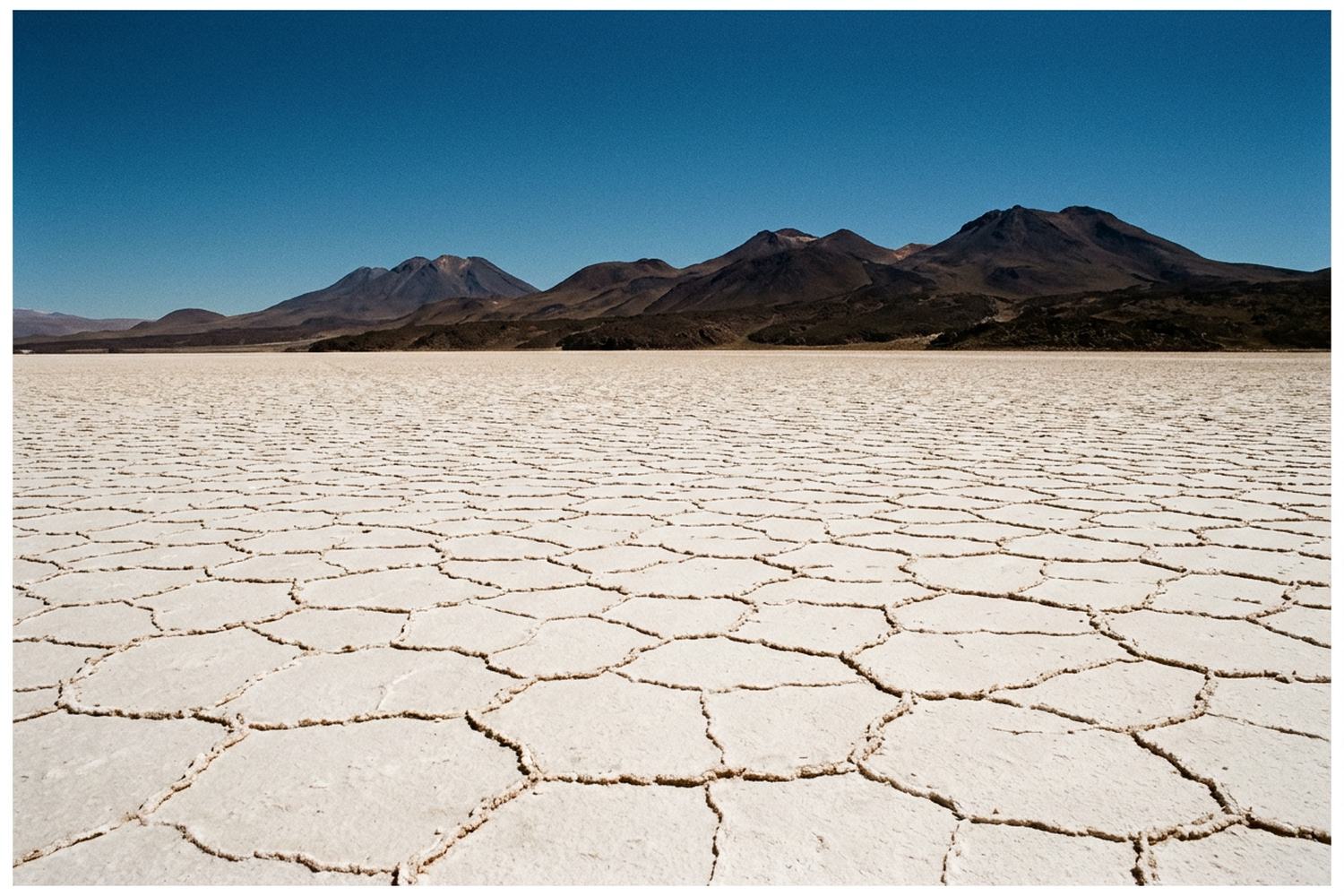 A white salt flat cracked into polygons running to dark volcanic cones under a deep blue sky