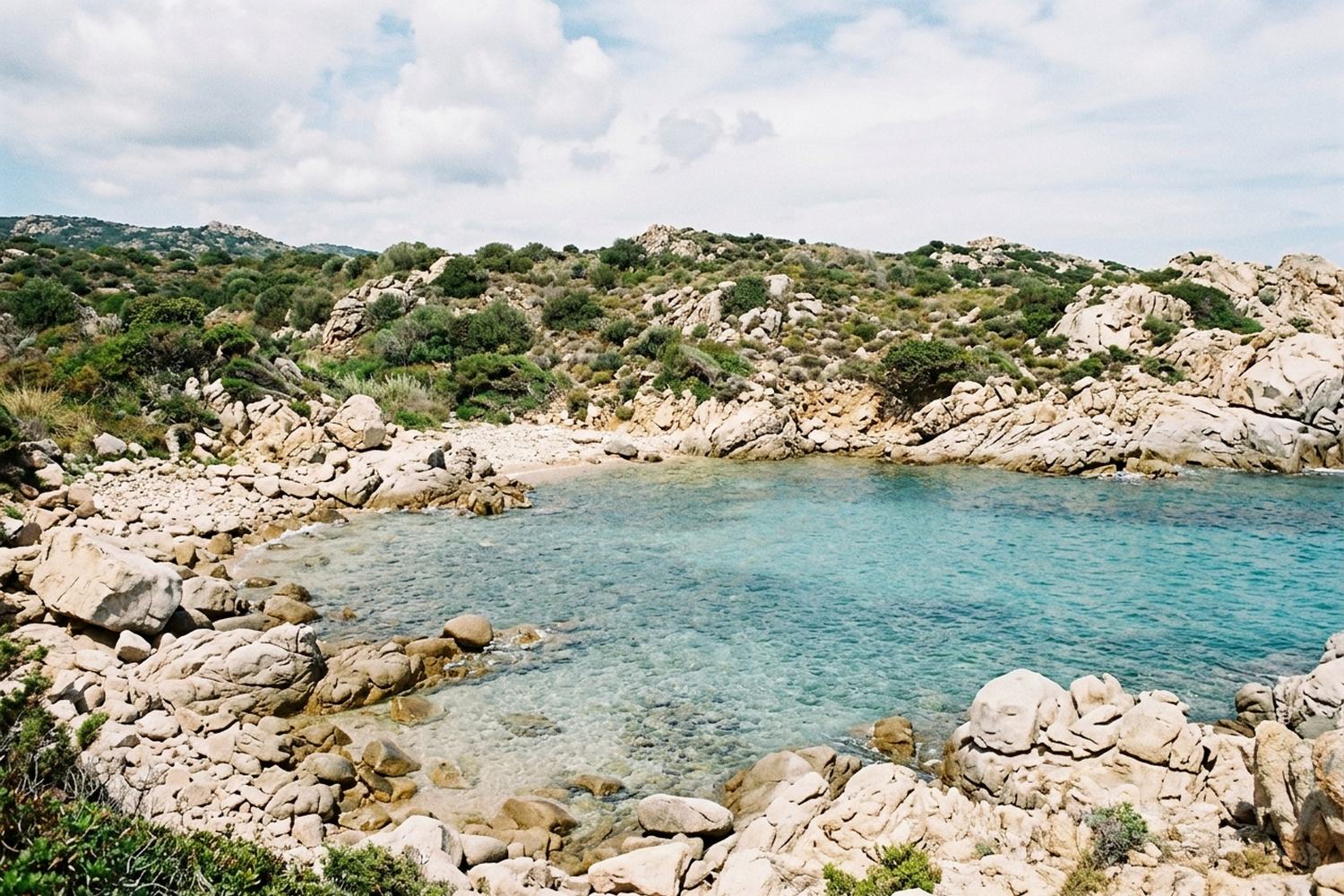 A small cove of pale granite boulders with clear turquoise water and low scrub